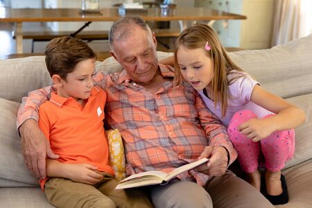 Front view of a senior Caucasian man at home in the living room sitting on the couch between his grandson and granddaughter, holding a book and reading togetherの写真素材