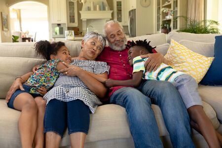 Front view of a senior mixed race couple at home in the living room, sitting on the couch with their young grandson and granddaughter, smiling at each other, relaxing and embracingの写真素材