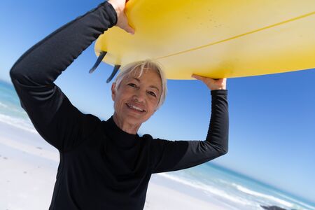 Senior Caucasian woman enjoying time at the beach, standing and holding a surfboard above her head