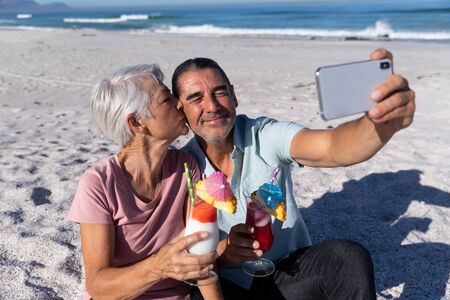 Senior Caucasian couple enjoying time at the beach, embracing, holding drinks and taking a selfie, a woman is kissing a man on his cheekの写真素材