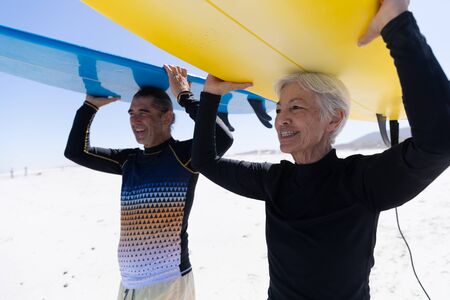 Senior Caucasian couple enjoying time at the beach, walking towards the sea and holding surfboards above their headsの写真素材