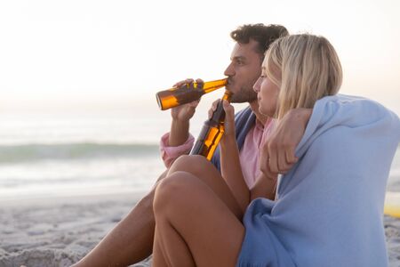 Caucasian couple enjoying time at the beach, sitting covered with a blanket, embracing and drinking beer, during a sunsetの写真素材