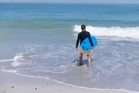 Senior Caucasian man enjoying time at the beach, walking towards the sea and holding a surfboard
