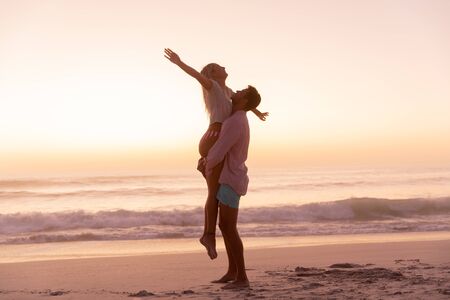 Caucasian couple enjoying time at the beach, a man is lifting a woman, a woman is widening her armsの写真素材