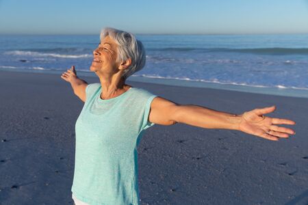 Senior Caucasian woman enjoying time at the beach on a sunny day, widening her arms and smiling with...