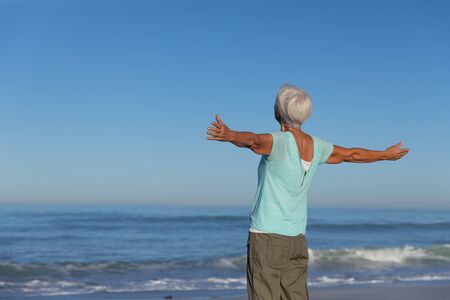 Senior Caucasian woman enjoying time at the beach on a sunny day, widening her arms and smiling with sea in the backgroundの写真素材
