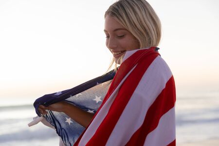 Caucasian woman enjoying time at the beach during a sunset, covering herself with an US flag with blue sea in the backgroundの写真素材