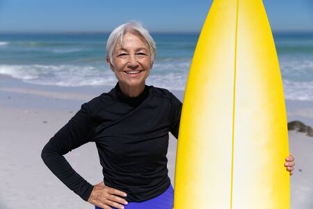 Senior Caucasian woman enjoying time at the beach on a sunny day, standing and holding a surfboard with sea in the backgroundの写真素材