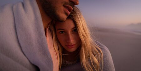 Caucasian couple enjoying time at the beach during a pretty sunset, embracing and covering...