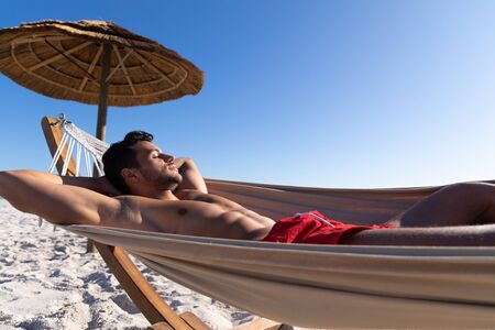 Caucasian man enjoying time at the beach on a sunny day, lying on a hammock and sunbathing with sea in the backgroundの写真素材