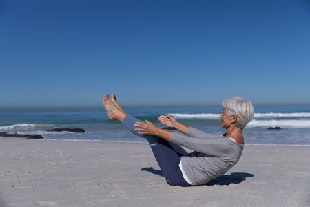 Senior Caucasian woman enjoying time at the beach on a sunny day, sitting on sand and practising yoga with sea in the backgroundの写真素材