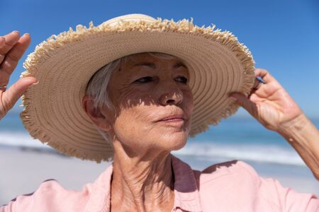 Senior Caucasian woman enjoying time at the beach on a sunny day, wearing a hat and smiling with sea in the backgroundの写真素材