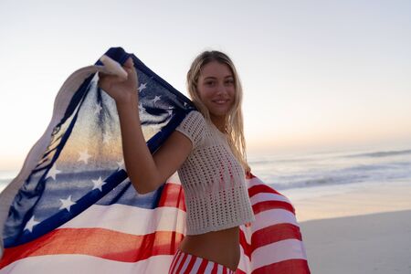 Caucasian woman enjoying time at the beach during a sunset, holding and waving an US flag with blue...