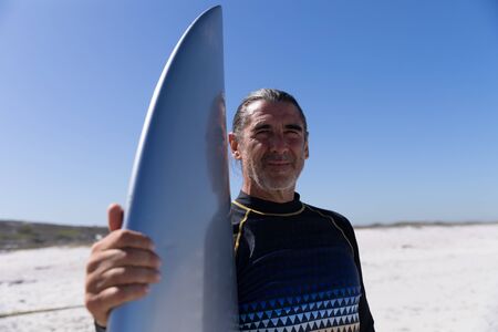 Senior Caucasian man enjoying time at the beach on a sunny day, standing and holding a surfboard...