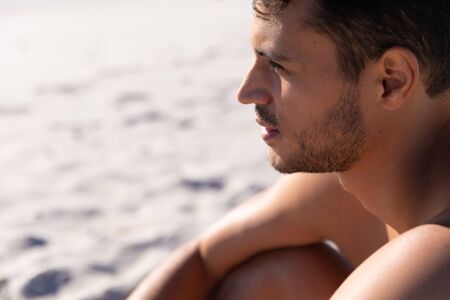 Caucasian man enjoying time at the beach on a sunny day, sitting and looking away with sand in the backgroundの写真素材