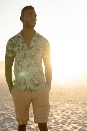 An African American man enjoying free time on beach on a sunny day, standing on sand with sun shining behind him, wearing a Hawaiian shirt. Relaxing summer vacation.の写真素材