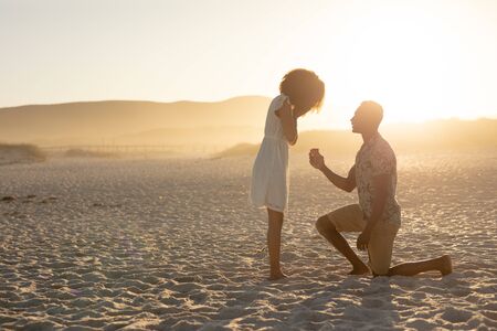 Mixed race couple enjoying free time on beach on a sunny day together, man proposing to the woman, kneeling on one knee on the sand. Relaxing summer vacation.の写真素材
