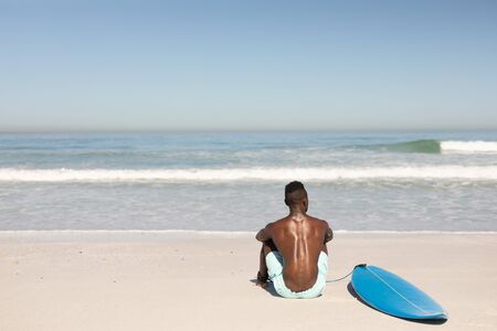 A rear view of an attractive African American man enjoying free time on beach on a sunny day, smiling, having fun, surfing, sitting on the beach, sun shining on him.の写真素材