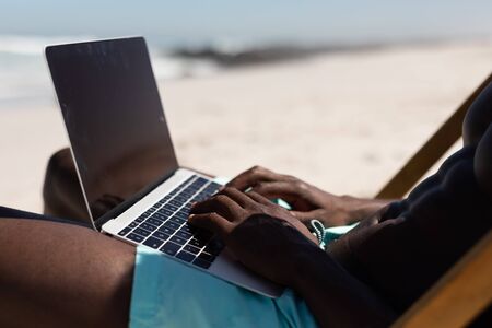 An attractive African American man enjoying free time on beach on a sunny day, sitting in a deck chair, using his laptop.の写真素材