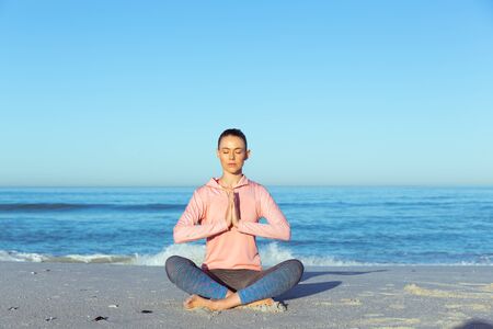 A Caucasian woman wearing sports clothes, enjoying time at the beach on a sunny day, practicing yoga, with eyes closed and hands in a prayer positionの写真素材