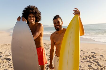 Two mixed race men enjoying their time at the beach with their friends on a sunny day, holding surfboards, looking at the camera and smilingの写真素材