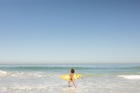 an attractive mixed race woman enjoying free time on beach on a sunny day, wearing a swimsuit, walking on the sand, surfing, carrying her surfboard, sun shining on her face.の写真素材