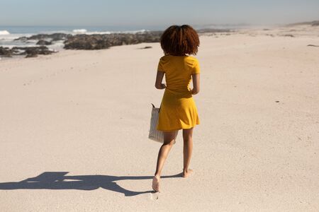 A rear view of an attractive mixed race woman enjoying free time on beach on a sunny day, wearing a yellow dress, walking on the sand, sun shining on her face.の写真素材