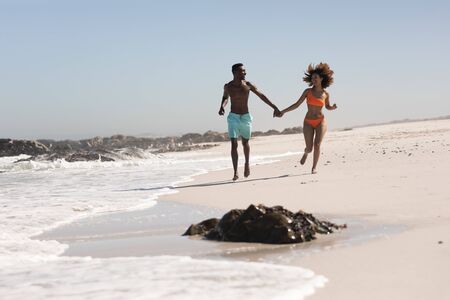 A mixed race couple enjoying free time on beach on a sunny day together, running and holding each others hands with sun shining on their faces.の写真素材