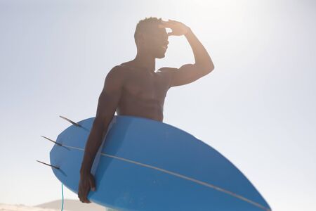 A happy, attractive African American man enjoying free time on beach on a sunny day, having fun, surfing, holding a surfboard, sun shining on his face.の写真素材