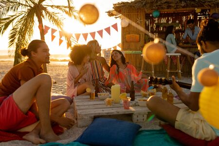 A multi-ethnic group of people enjoying their time at a beach with their friends during a sunset, sitting on sand, playing guitar and smilingの写真素材