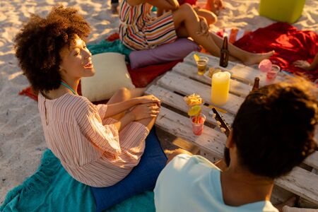 A multi-ethnic group of people enjoying their time at a beach with their friends during a sunset, sitting on sand, playing guitar and smilingの写真素材