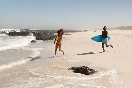A mixed race couple enjoying free time on beach on a sunny day together, surfing, running with their surfboards, having fun with sun shining on their faces.の写真素材
