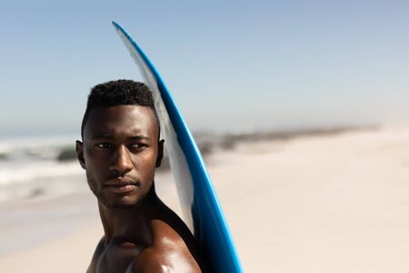 An attractive African American man enjoying free time on beach on a sunny day, having fun, standing with his surfboard, sun shining on him.の写真素材