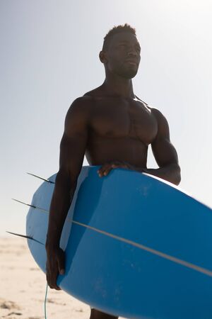 An attractive African American man enjoying free time on beach on a sunny day, having fun, surfing, holding a surfboard, sun shining behind him.の写真素材