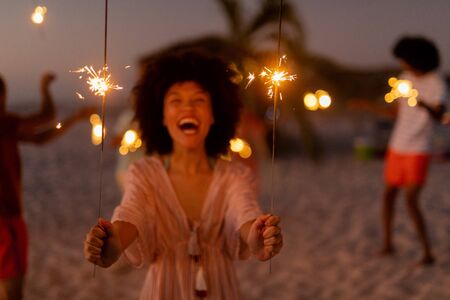 Mixed race woman enjoying her time at the beach with her friends during sunset, holding sparklers, looking at the camera and smiling with her friends in the backgroundの写真素材