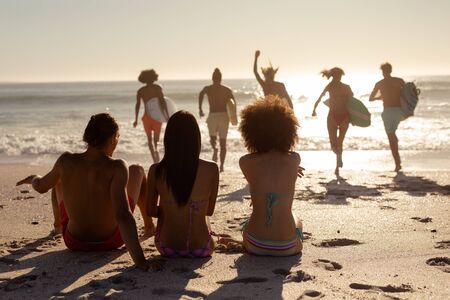 Rear view of a multi-ethnic group of three female friends on holiday sitting on a beach and watching their friends running into the sea holding surfboards as the sun goes downの写真素材
