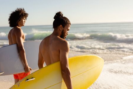 Close up of two Caucasian and mixed race male friends on holiday standing on a sunny beach holding...
