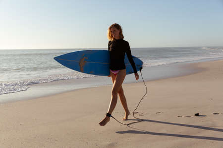 Attractive blonde Caucasian woman enjoying time at the beach on a sunny day, holding surfboard and...