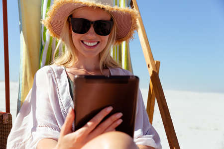 Attractive blonde Caucasian woman enjoying time at the beach on a sunny day, sitting on deck chair,...