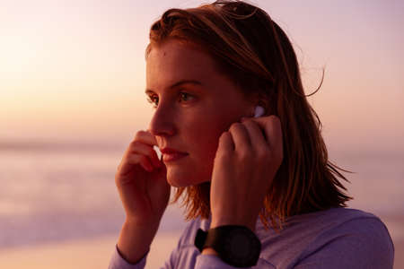 Attractive blonde Caucasian woman enjoying time at the beach at sundown, wearing earphones, with...