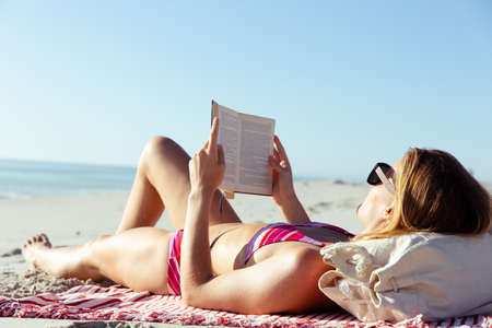 Attractive blonde Caucasian woman enjoying time at the beach on a sunny day, lying on a towel and reading a book, with blue sky and sea in the background. Summer tropical beach vacation.の写真素材