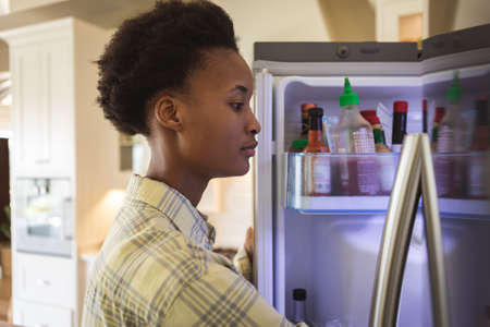 Mixed race woman spending time at home, standing in her kitchen and looking into a refrigerator. Self isolating and social distancing in quarantine lockdown during coronavirus covid 19 epidemic.の写真素材