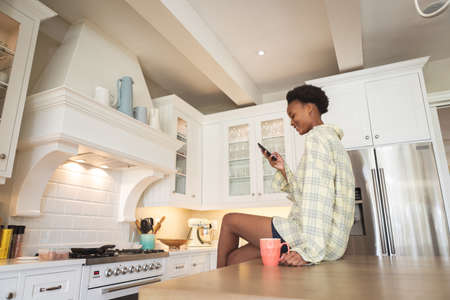 Mixed race woman spending time at home, sitting in her kitchen and using a smartphone. Self isolating and social distancing in quarantine lockdown during coronavirus covid 19 epidemic.の写真素材