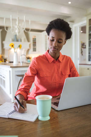 Mixed race woman spending time at home, sitting in her kitchen and using her laptop computer. Self isolating and social distancing in quarantine lockdown during coronavirus covid 19 epidemic.の写真素材