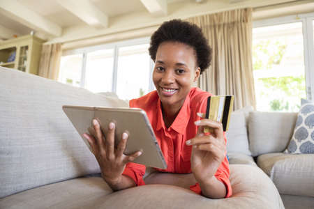 Mixed race woman spending time at home, lying on couch, using tablet while online shopping. Self...