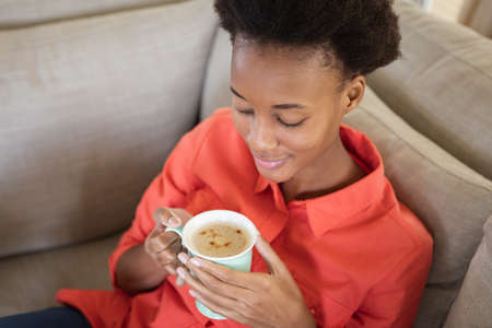 Mixed race woman spending time at home, sitting on couch and holding a mug of coffee. Self isolating and social distancing in quarantine lockdown during coronavirus covid 19 epidemic.の写真素材