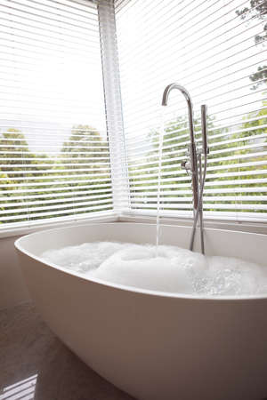 Modern bathroom interior showing free standing bath full of water and foam and water running from...