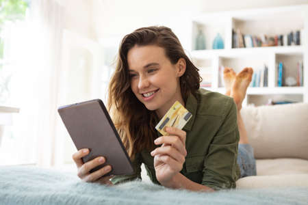 Caucasian woman spending time at home, sitting on sofa, using tablet, holding credit card. Lifestyle...