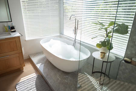 Modern bathroom interior showing free standing bath with large windows showing green trees and plants outside, with white bathtub and silver water tap, grey shower and a table with a plant.の写真素材