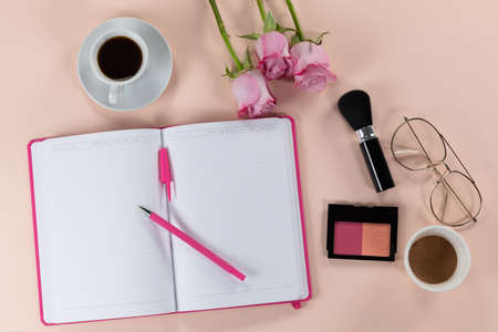 Top view of a notebook, a brush, a blush, powder, a cup of coffee and glasses, with pink roses arranged on a plain pink backgroundの写真素材